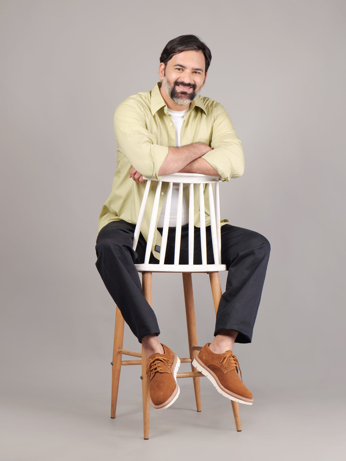 Man sitting on a white stool wearing a light green shirt, black pants, and brown shoes against a beige background