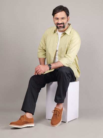 Male model wearing brown Blucher Suede shoes, seated on a white cube in a studio setting
