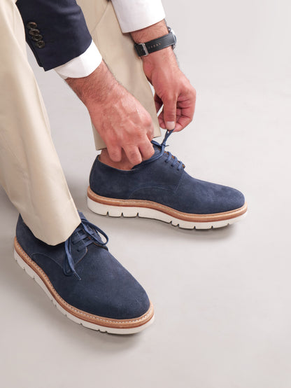 Navy blue Blucher Suede shoes while a model ties the laces against a beige background
