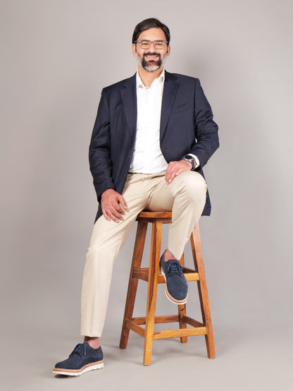 Male model wearing navy blue Blucher Suede shoes while seated on a stool against a beige background